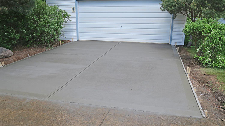 Freshly poured concrete driveway in front of a garage, surrounded by greenery in a residential yard setting